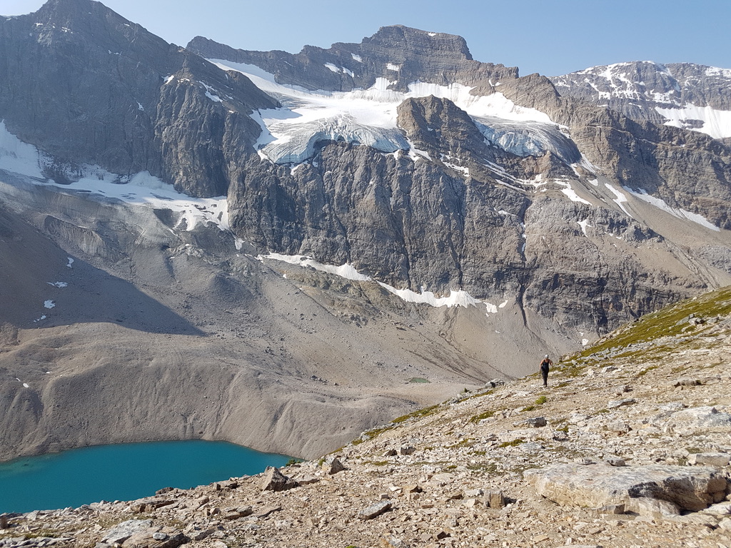 A hiker on a rocky slope with glacier-covered mountains behind and a blue-green tarn below.